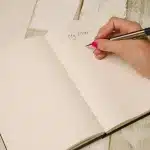 Manos anónimas hojeando un manuscrito sobre una mesa de café, junto a un bolígrafo y libros desenfocados