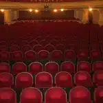 Espectáculo teatral con iluminación colorida, performers en escena y público de espaldas observando